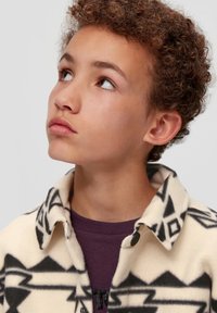 Fleece-Jacke in Creme mit schwarzen geometrischen Mustern, mit einem geknöpften Kragen und übereinander getragen über einem dunkelvioletten Hemd. Strukturierter Stoff.