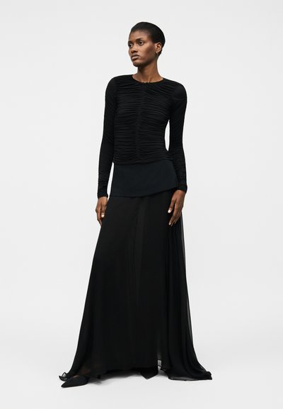 Kvinde står iført en lang sort rynket bluse med lange ærmer og et flydende sort gulvlængde nederdel med sorte spidse flade sko.