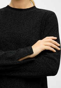Persoon in een lange mouwen zwarte textuurtop, met één arm gekruist en de hand rustend op de tegenoverliggende arm tegen een effen achtergrond.