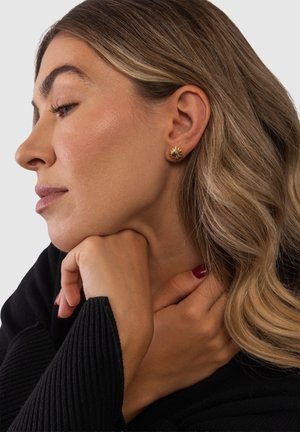 Frau mit welligem blondem Haar, die das Kinn auf die Hand stützt, trägt einen schwarzen Pullover und eine kleine goldene Sonnenförmige Steckerohrring.