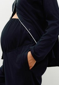Pregnant person wearing loose black pants and black button-up top with white piping, hand resting inside pants pocket.