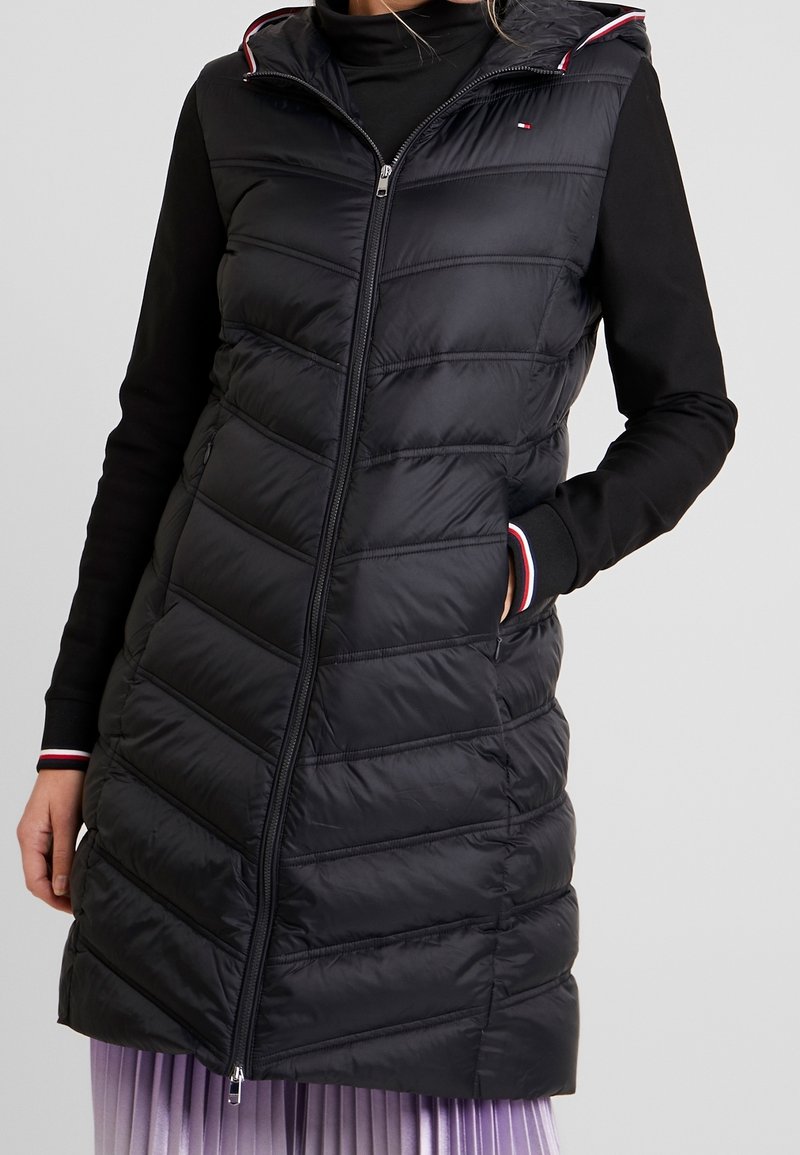 Zwarte gewatteerde mouwloze vest met een capuchon, met een rits aan de voorkant, horizontale stiksels en rode en witte accenten langs de randen.