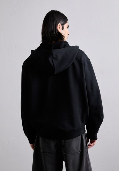 Črna oversize kapuca iz mehkega materiala, z velikim pokrovom, spušenimi rameni in rebrastimi manšetami. Oblečena z temnimi, ohlapnimi hlačami.