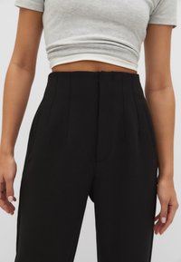 Pantaloni neri a vita alta con pieghe frontali, tasche laterali e una texture liscia, abbinati a un top grigio corto.