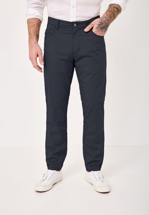 Mann trägt dunkelblaue Hose, weiße Sneakers und ein weißes Hemd mit Knöpfen, eine Hand in der Tasche, zeigt Unterarm-Tattoos.