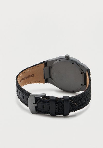 Πίσω όψη ενός μαύρου ρολογιού χειρός με υφασμάτινο λουρί με υφή, μεταλλική αγκράφα και οκταγωνική κάσα που δείχνει έξι βίδες σε απλή φόντο.