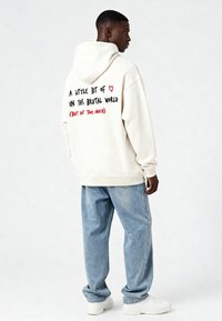 Kreme krūze ar kanguru kabatu un melnu un sarkanu tekstu aizmugurē; kombinēta ar gaiši zilu džinsu un baltām sporta apaviem.