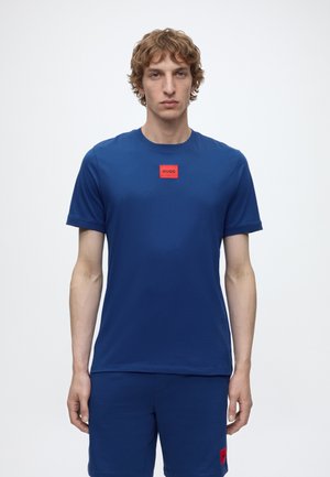 Jeune homme portant un ensemble t-shirt et short bleu avec des écussons rouges "HUGO", debout devant un fond blanc uni.