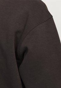 Material de sudadera marrón oscuro con una textura suave y costuras visibles. Presenta un cuello redondo y mangas raglán.