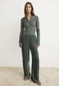 Zeleni baršunasti kombinezon s dugim rukavima, dubljim V-izrezom i opuštenom strukom. Ima široke nogavice i mekan, glatki tekstur.