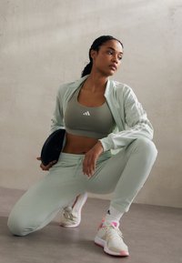 Light green athletic jacket and matching pants with white stripes. Olive sports bra with white logo. Beige sneakers with pink accents and white socks.