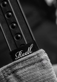 Black strap with small circular studs, featuring a gray fabric end with "Reell" embroidered in white cursive on a black background.