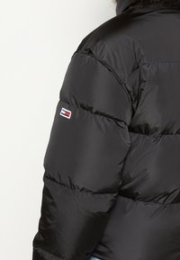 Doudoune noire avec un design matelassé, fabriquée en matériau brillant, avec un petit logo brodé sur la manche gauche.