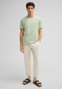 Hellgrünes Baumwoll-T-Shirt, lockere Passform, kurze Ärmel. Beigefarbene Leinenhose, am Knöchel umgeschlagen. Schwarze Sandalen mit zwei verstellbaren Riemen.