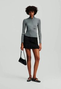 Top cinza com nervuras, com fecho zip e mangas longas, combinado com uma saia preta com babados e sapatos slip-on pretos com tachas.