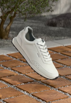 Fehér, sima bőrből készült sportcipő alacsony profilú, kerek orral és lapos fűzőkkel. Az oldalak perforáltak, fekete gumi talppal.