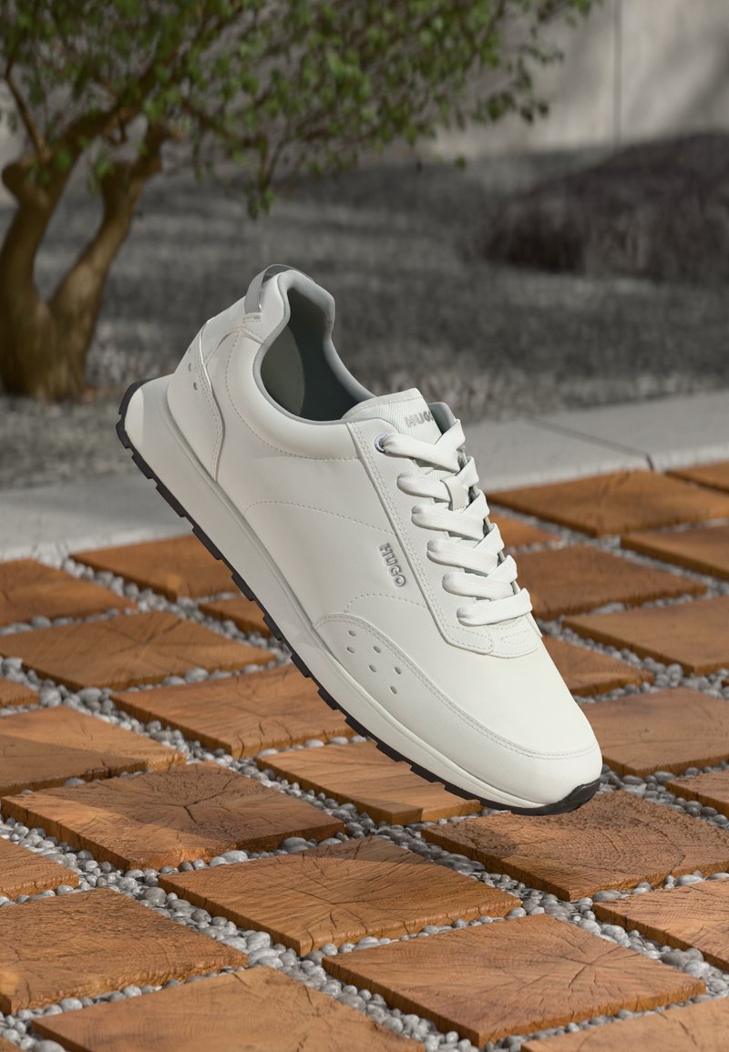 Valge tennisekreem, millel on sile nahk, paeltega disain, minimalistlikud aksessuaarid ja tekstureeritud kummist tald, riputatud puidust plaatide kohal.