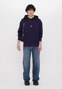 Navy blue hoodie with drawstrings and logo on chest, paired with light blue jeans and black shoes, worn with a small black crossbody bag.