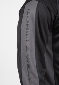 Zwarte sportjack met een gladde textuur en een contrasterende grijze streep, voorzien van de tekst "GORILLA WEAR" in zwarte letters langs de arm.