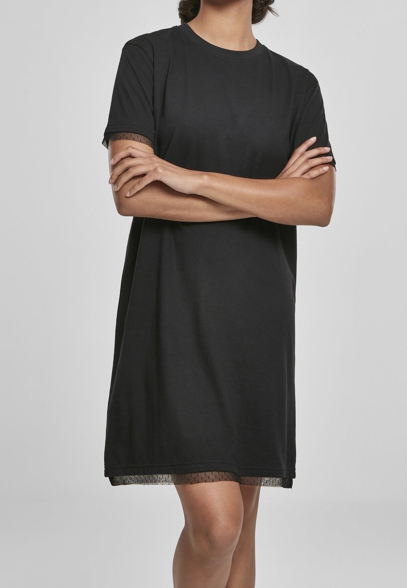 Personne portant une robe noire à manches courtes avec un bord en tulle à pois, debout les bras croisés devant un fond uni.