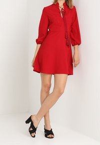 Červené šaty s límečkem, detailem ve tvaru klíčové dírky a šňůrkou na stažení. Dlouhé nabírané rukávy a rozšířená sukni. Nosit s černými sandály na podpatku.