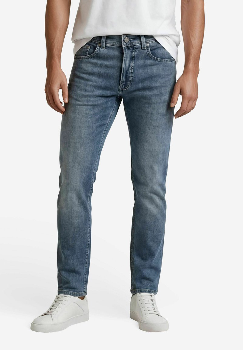 Mann trägt eng anliegende blaue Jeans, weiße Turnschuhe und ein weißes, eingestecktes T-Shirt und steht vor einem schlichten weißen Hintergrund.