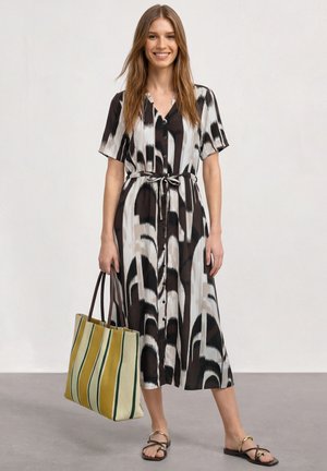 Femme en robe midi à motifs noir et blanc tenant un grand sac cabas rayé jaune et vert, portant des sandales noires et souriant.