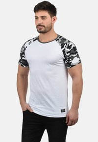Weißes T-Shirt mit kurzen Ärmeln und grauen Camouflage-Muster-Raglanärmeln sowie einem Rundhalsausschnitt. Ausgestattet mit einem kleinen schwarzen Etikett am Saum.