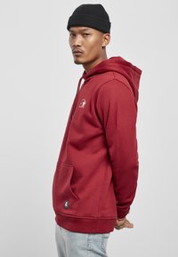 Rød hettegenser laget av mykt stoff, med lomme foran, logo brodert på brystet og ribbestrikkede mansjetter. Båret med lyse jeans.