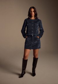 Veste en velours côtelé marine avec deux poches poitrine, jupe assortie et bottes noires montantes. Texture lisse, coupe ajustée et détails métalliques.