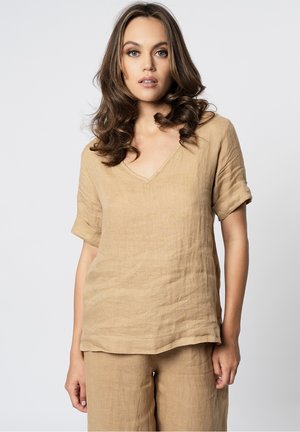 Donna con capelli castani ondulati che indossa una maglietta beige di lino a scollo a V e maniche corte e pantaloni abbinati, in piedi di fronte a uno sfondo chiaro e uniforme.