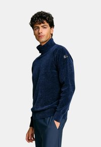 Maglione a collo alto blu navy in un tessuto morbido e testurizzato. Presenta una vestibilità rilassata e un accento con il marchio sulla manica sinistra.
