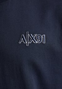 Tela de algodón azul marino con un logo bordado que presenta "A|X91" en hilo blanco, situado en el centro del área del pecho.