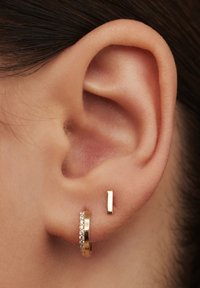 Goldfarbene Creolen mit Strasssteinen und ein minimalistischer goldfarbener quadratischer Stecker, der am Ohr positioniert ist. Glatte Textur und glänzende Oberfläche.