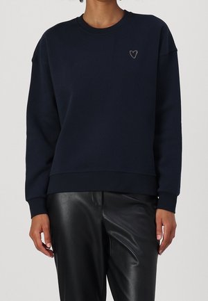 Mujer con sudadera azul marino con un pequeño corazón bordado en el pecho y pantalones de cuero negros, de pie frente a un fondo liso.