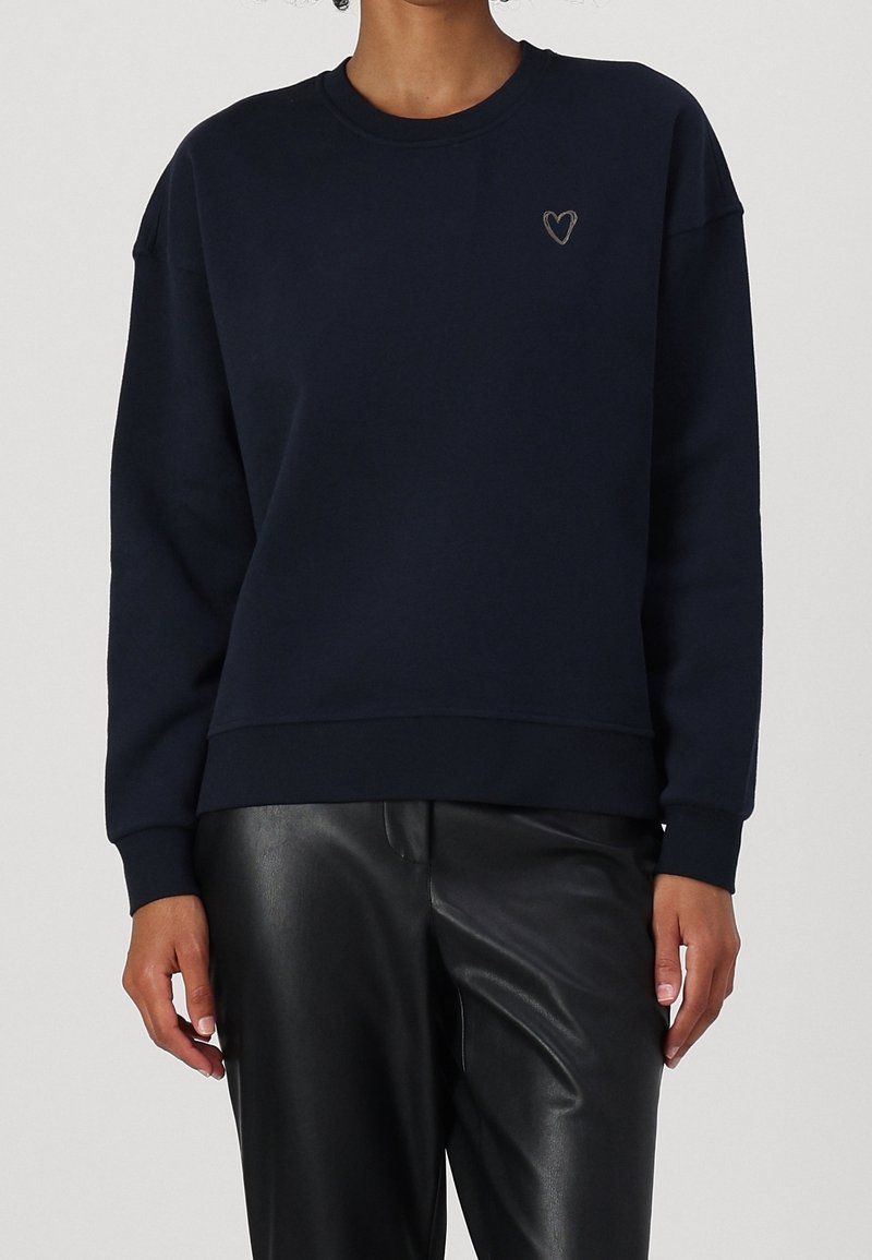 Femme portant un sweat-shirt bleu marine avec un petit cœur brodé sur la poitrine et un pantalon en cuir noir, debout devant un fond uni.
