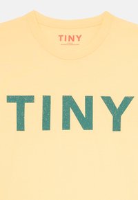 T-shirt en coton jaune avec de grandes lettres texturées en bleu canard orthographiant "TINY" sur le devant, avec un col rond et un tissu doux.