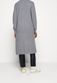 Cardigan long gris côtelé à manches longues, associé à un pantalon en cuir noir et des baskets blanches avec des accents jaunes.