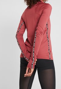 Langärmliges Sportoberteil in Rostrot, mit figurbetontem Design, glatter Textur und seitlichen Streifen mit schwarzer Textmarkierung.