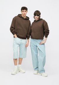 Braune Kapuzenpullover mit Fronttaschen, kombiniert mit hellblauen Jeansshorts und Hosen. Beide Modelle tragen weiße Schuhe mit einem klobigen Design.