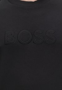 Tissu noir avec le mot BOSS brodé en grandes lettres noires en relief sur la zone de la poitrine d'un vêtement.