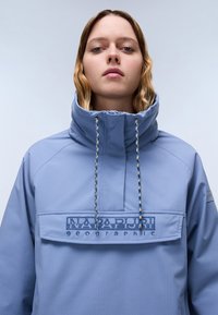 Lyseblå genserjakke med høy krage, med en fremre lomme og brodert logo. Strukturert stoff og kontrasterende snorer.