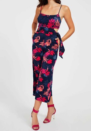 Femme portant une combinaison bleu marine sans manches avec un motif floral rouge et rose et des sandales rouges à talons assorties.