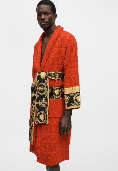 Άνδρας που φοράει κόκκινο ρόμπα Versace με ανάγλυφο λογότυπο, μαύρη και χρυσή ζώνη με σχέδιο και μανσέτες, στέκεται μπροστά σε απλό φωτεινό φόντο.