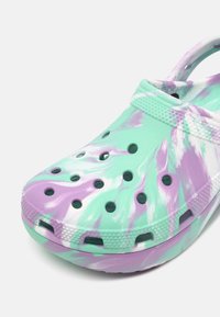 Sabot tie-dye vert clair et violet avec des trous de ventilation et une bride de talon texturée sur fond blanc.