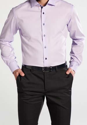Mężczyzna ubrany w jasnofioletową koszulę wizytową z ciemnymi guzikami i czarne spodnie wizytowe w kratę, z rękami w kieszeniach, stojący na jednolitym tle.