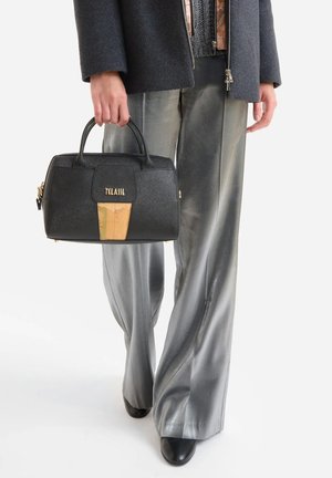 Persona che indossa pantaloni argento a gamba larga e giacca scura, che tiene una borsa nera strutturata con logo dorato e dettaglio mappa.