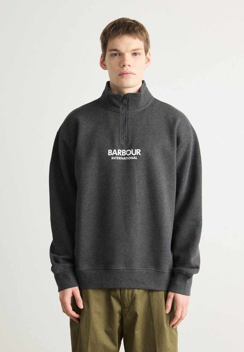 Tmavě šedá mikina na zip vyrobená z měkkého materiálu; má vysoký límec, spadlá ramena a bílé logo "BARBOUR INTERNATIONAL" na hrudi.