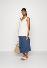 Top de maternidade em tecido bege claro, com decote em V; combinado com uma saia de ganga longa. Usando sandálias de deslizar bejes, segurando uma bolsa de mão em tom castanho.