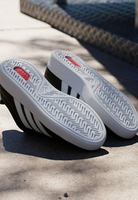 Paire de baskets noires et blanches reposant sur le côté, montrant des semelles blanches détaillées avec des accents rouges sur une surface en béton.